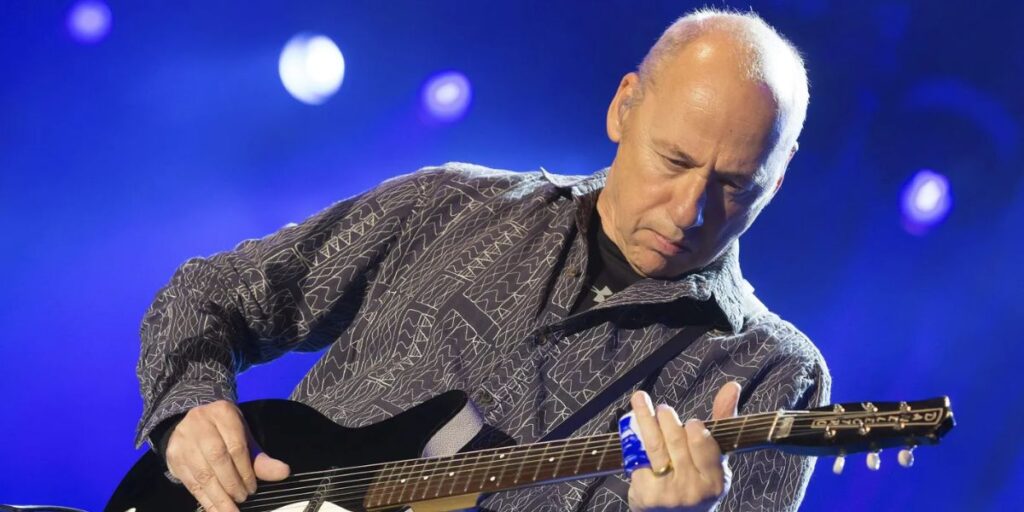 Mark Knopfler performing live on stage playing his signature fingerstyle guitar in 2025
